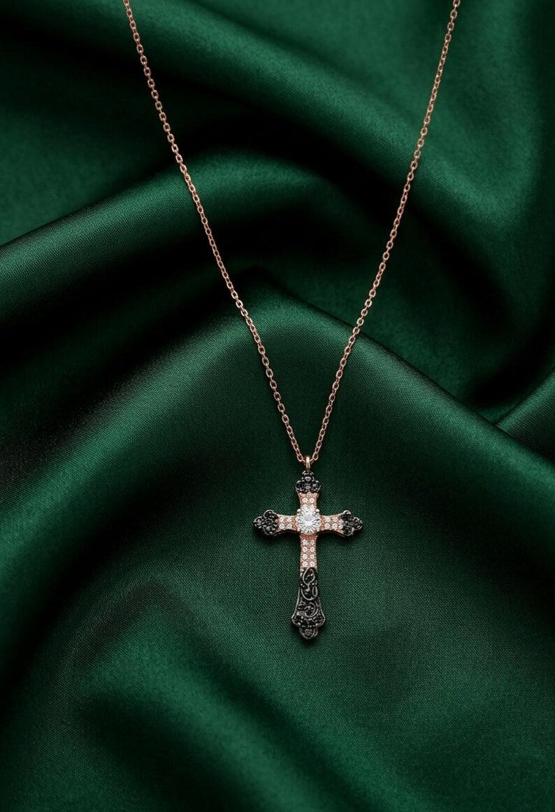 A rose gold cross pendant necklace with black and white gemstone accents, displayed on a dark green satin fabric background.