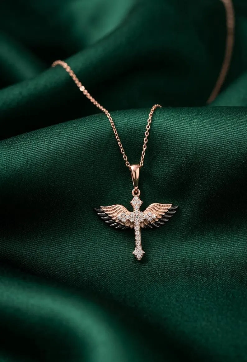 A rose gold winged cross pendant necklace adorned with small clear crystals, resting on draped dark green satin fabric.