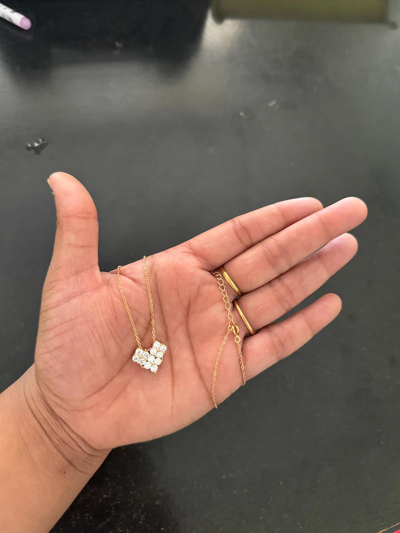 A gold chain necklace with a small cluster of clear stones pendant, held in a person's open palm. The person is also wearing a gold ring on their finger, against a dark gray tabletop background.