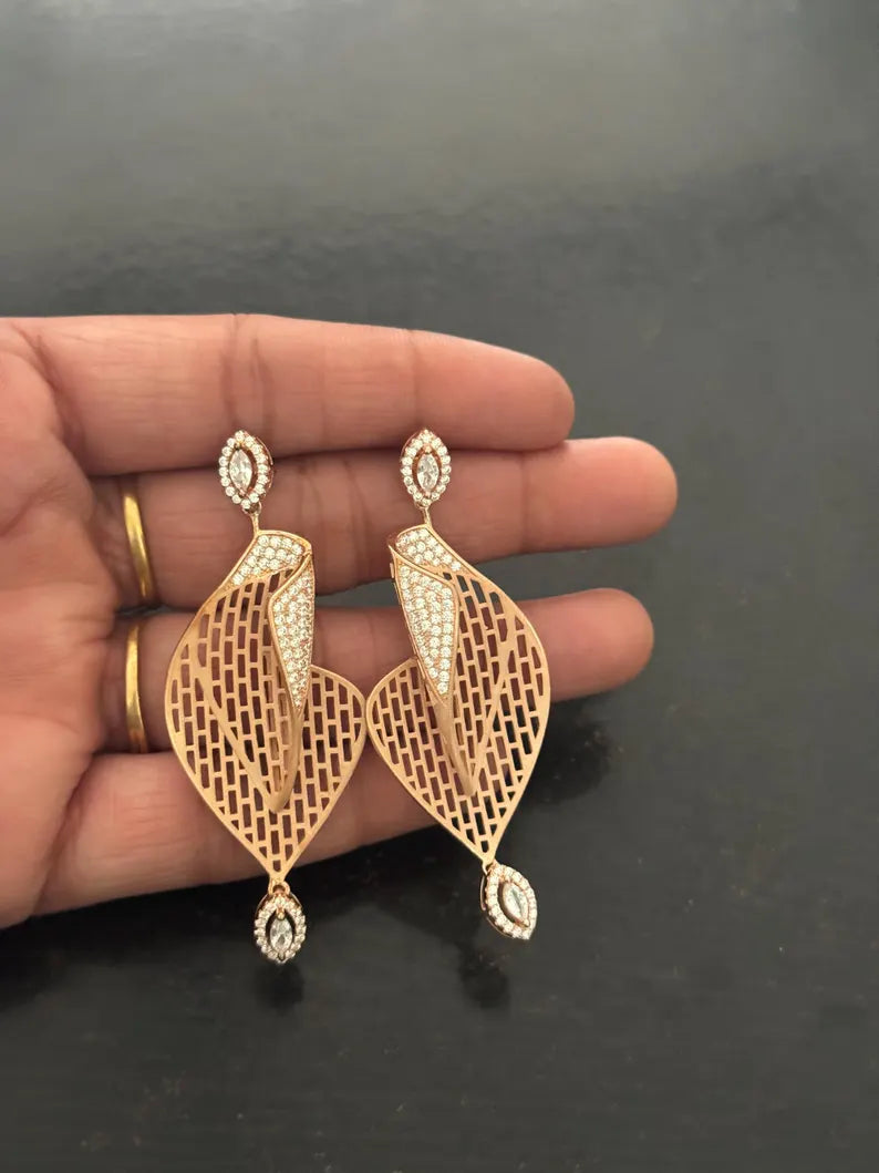 A pair of gold-toned dangle earrings with intricate filigree cutouts and pavé-set clear stones, held in a person's hand against a dark gray background.