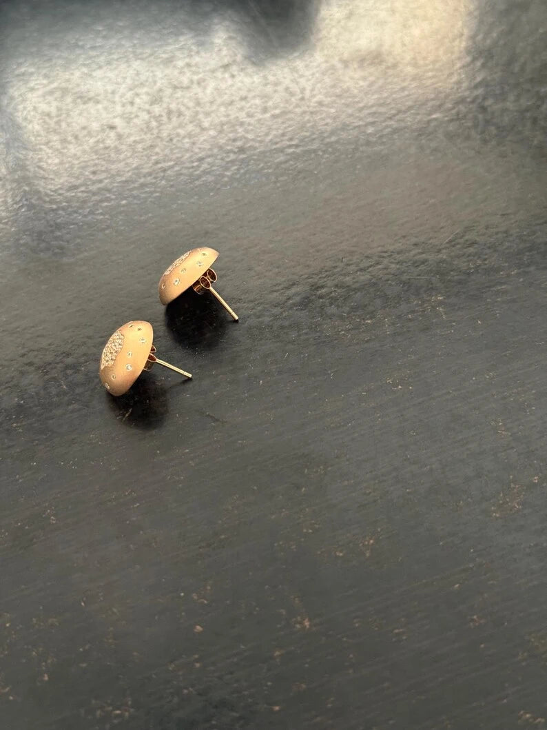 A pair of small dome-shaped stud earrings with a warm metallic finish, shown on a dark textured surface.
