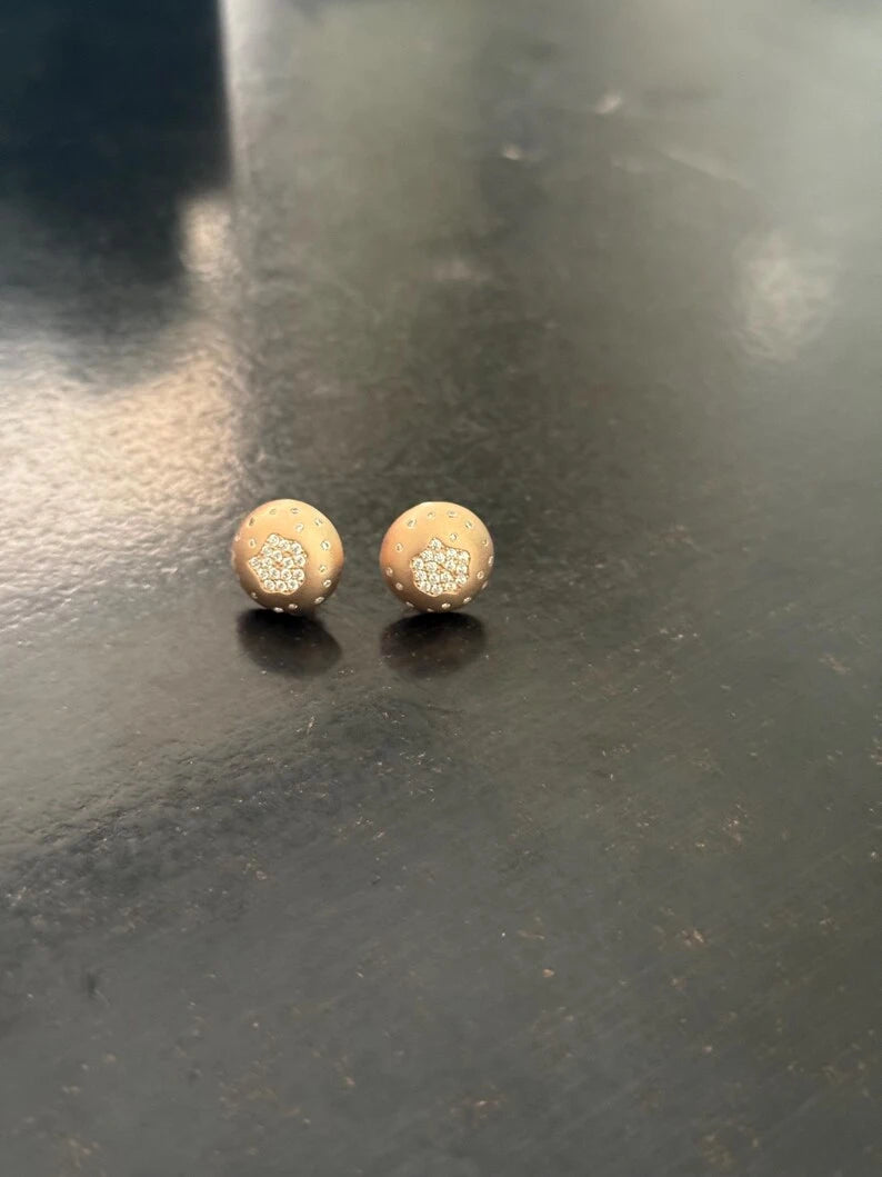 A pair of small round stud earrings with a textured gold finish and a small clear crystal heart detail, resting on a dark reflective surface.