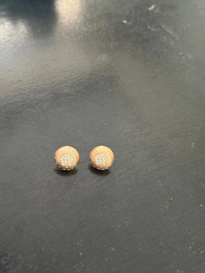 A pair of small round stud earrings with a textured beige center and gold-toned edges, resting on a dark gray surface.