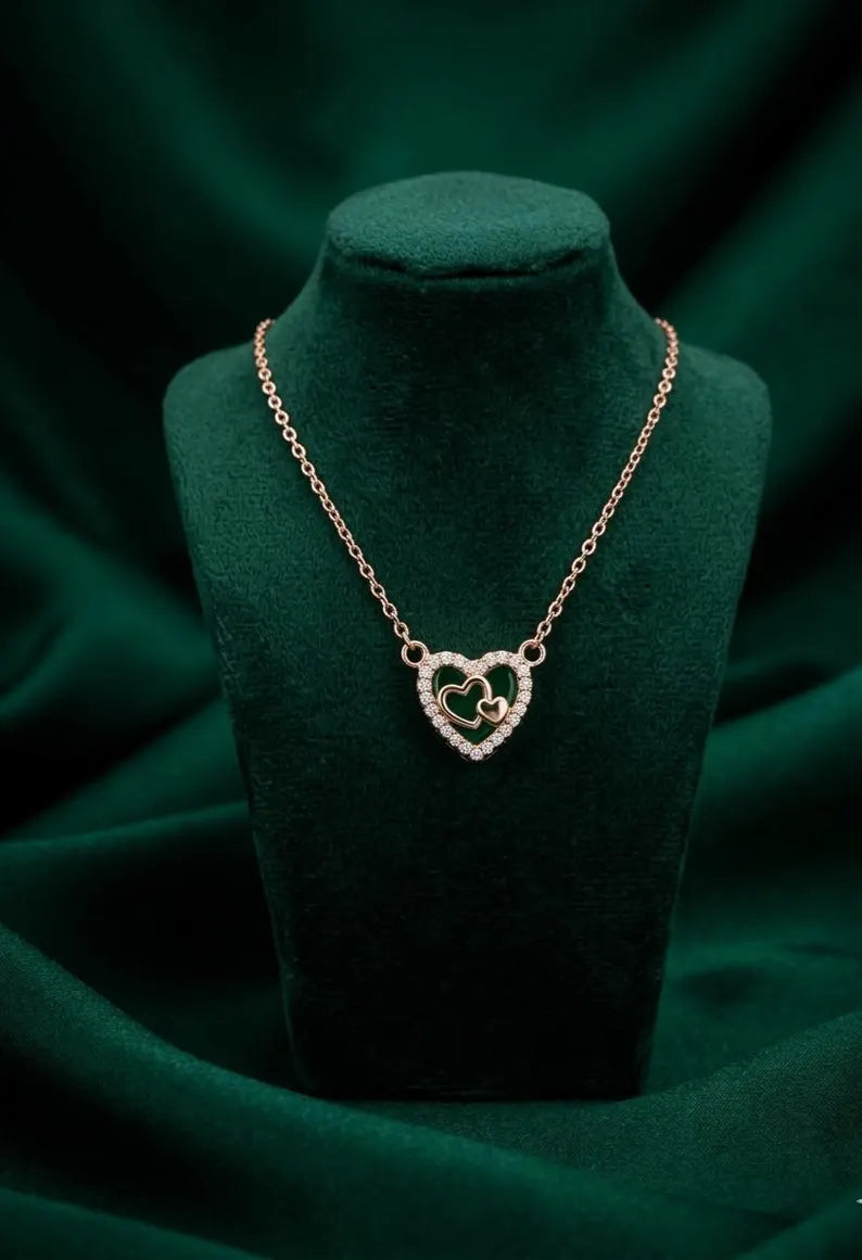 A heart-shaped pendant necklace with a gold-tone chain and double-heart pendant, displayed on a dark green velvet jewelry bust against a draped dark green fabric background.