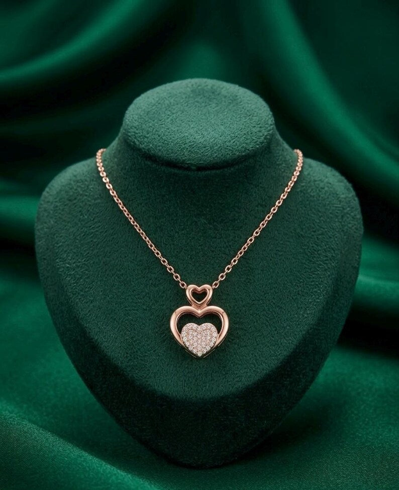 A rose gold double-heart pendant necklace with one heart featuring pavé diamonds, displayed on a dark green velvet jewelry bust against a matching green fabric background.