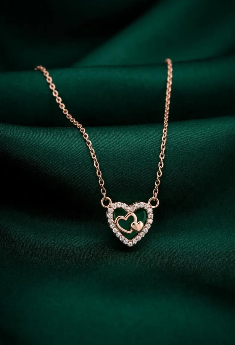 A rose gold double-heart pendant necklace with a sparkling crystal halo and a small gold heart in the center, resting on a draped dark green satin fabric.