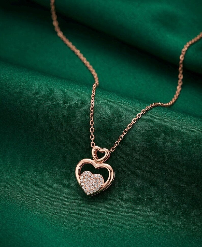 A rose gold necklace with a double-heart pendant, featuring a pavé-set clear heart in the center and an open heart outline above it, resting on a draped dark green satin fabric.