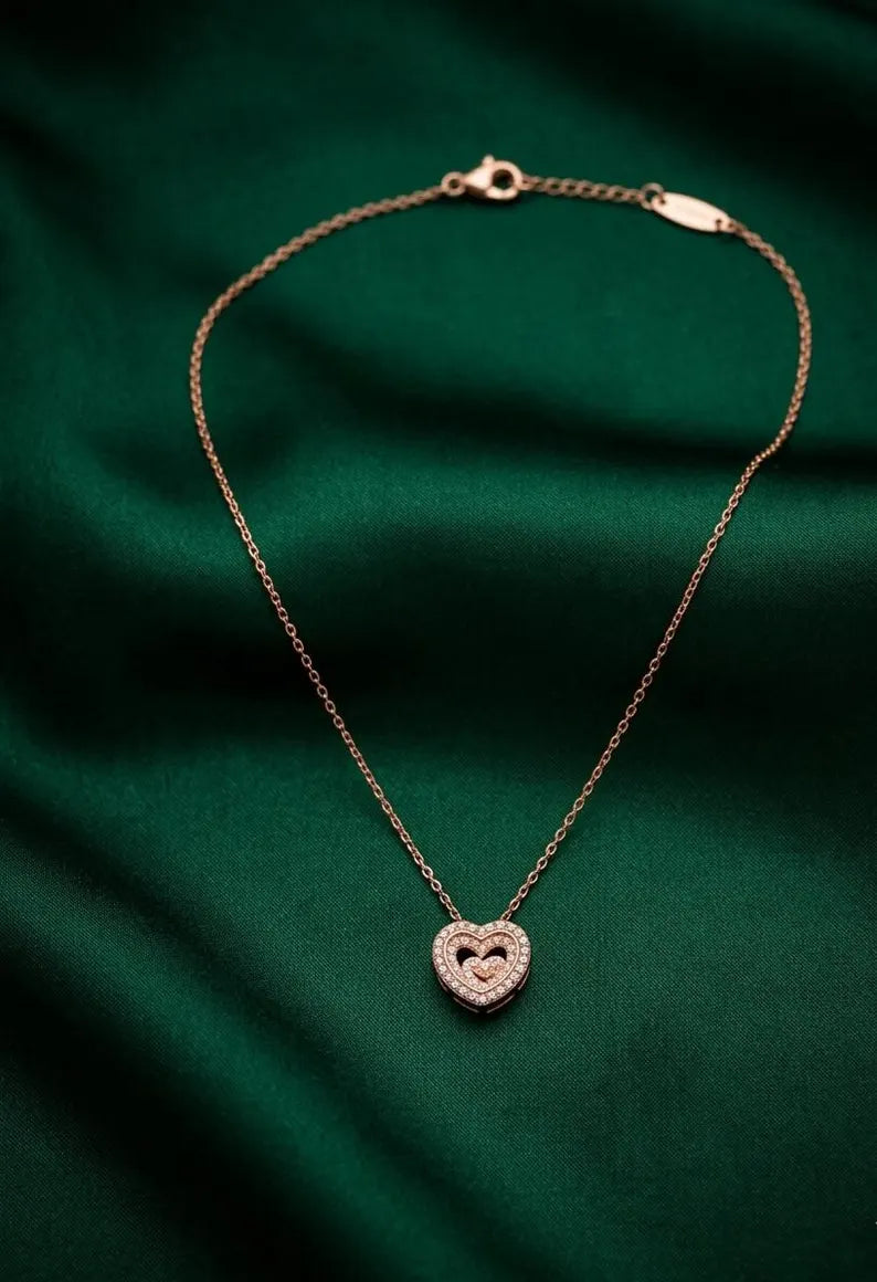 A delicate rose gold chain necklace with a heart-shaped pendant, displayed on a draped dark green satin fabric background.
