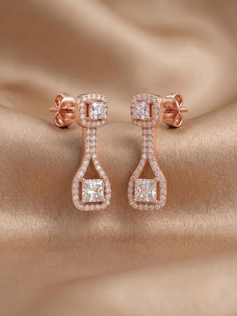 Rose gold drop earrings with princess-cut and round diamonds, displayed on a draped beige satin fabric background.