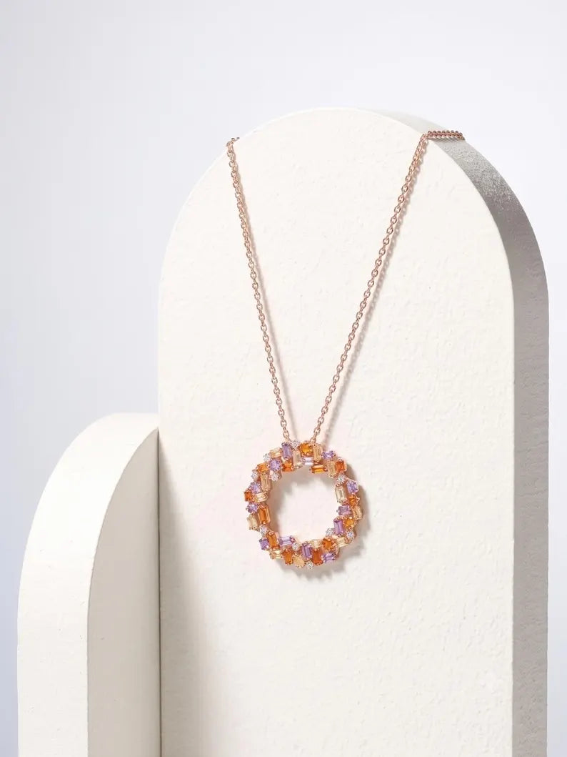 A delicate rose gold chain necklace with a circular pendant made of small amber and lavender faceted crystals, displayed on a white arched stand against a light gray background.