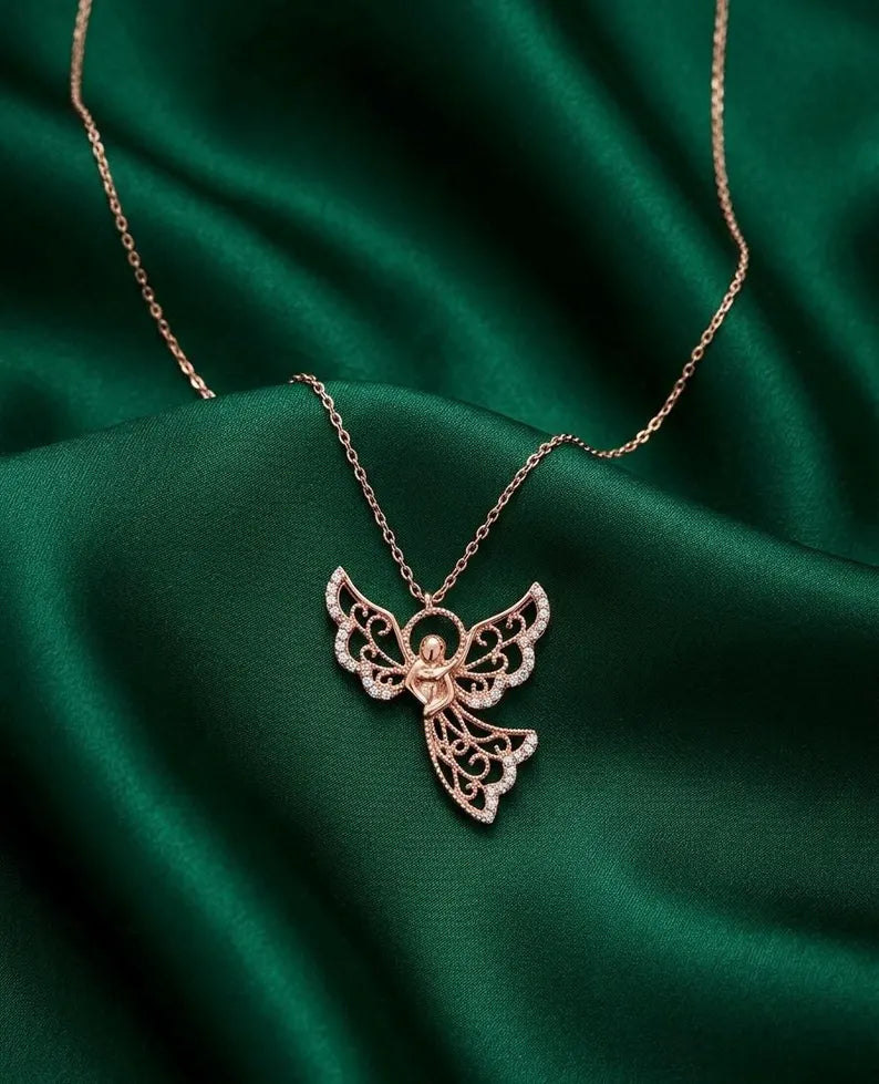 A rose gold angel pendant necklace with delicate filigree wings and small clear stones, displayed on draped emerald green satin fabric.