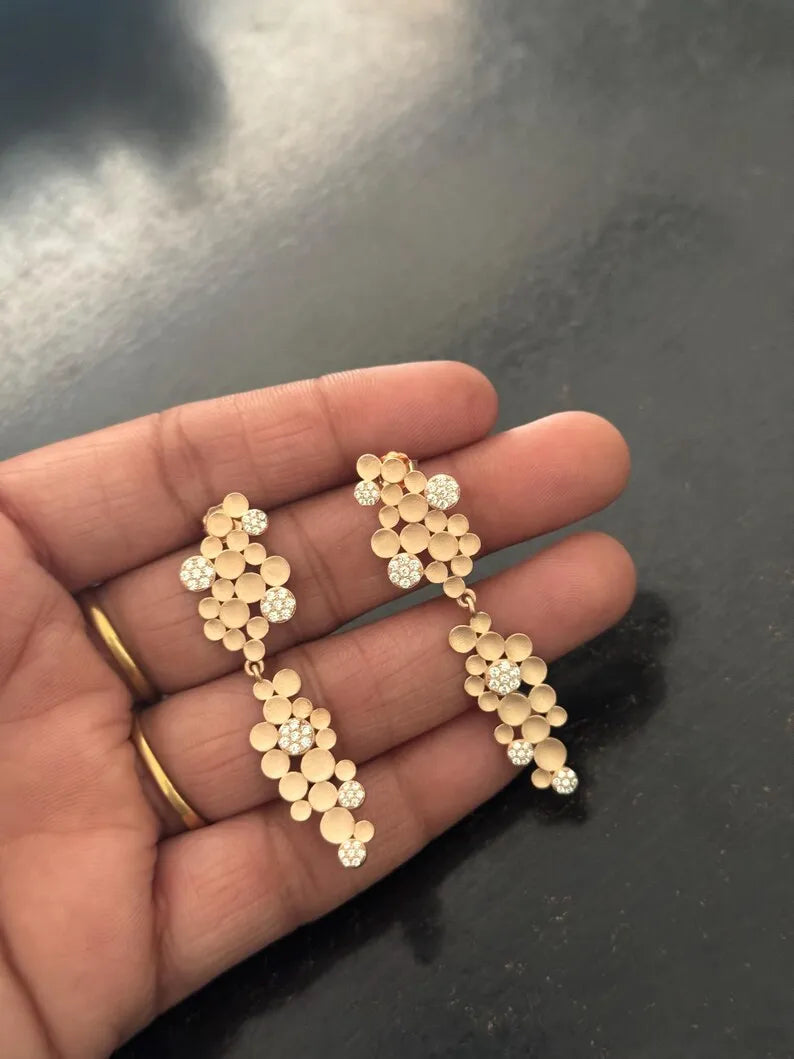 A pair of gold dangle earrings with clustered flower-like discs and small clear stones, shown resting on a person's hand against a dark reflective surface.