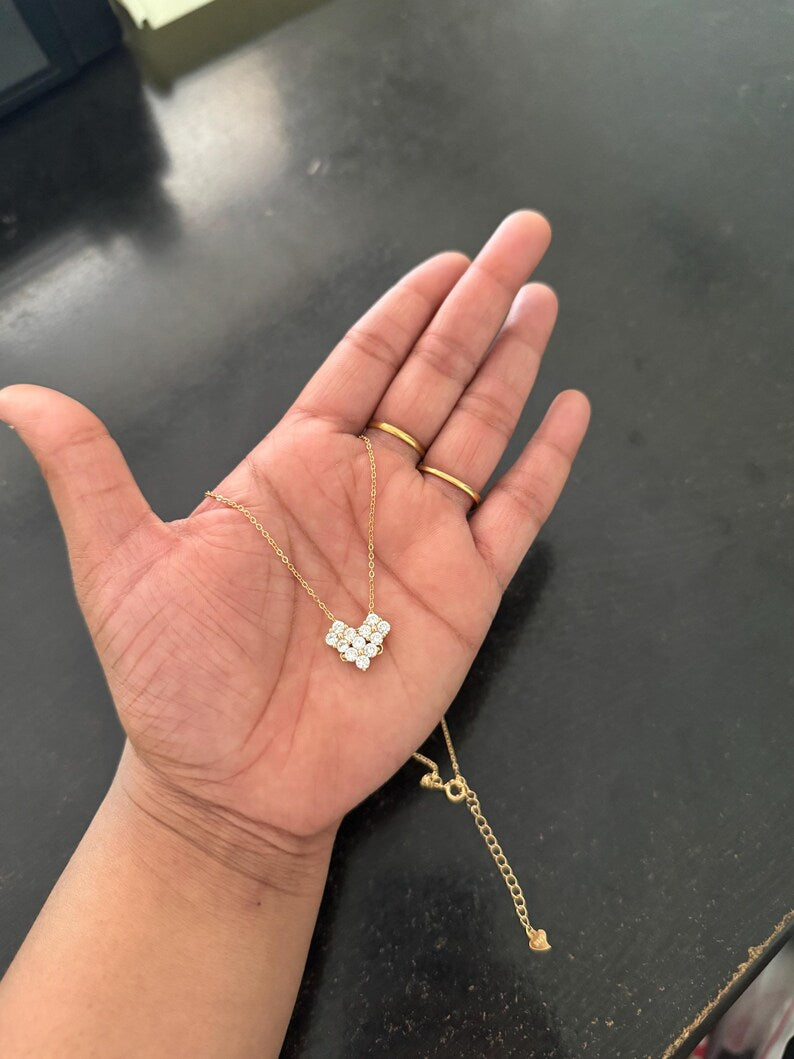 A gold chain necklace with a small white and clear stone heart-shaped pendant is draped over an open hand. The person holding the necklace wears two simple gold rings on their fingers, and the background is a dark, reflective surface.