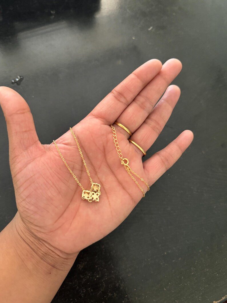 A gold necklace with a small geometric pendant is draped across an open hand, alongside a simple gold ring, against a dark gray surface.