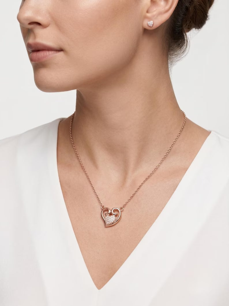 A close-up of a woman wearing a delicate rose gold heart pendant necklace with a pavé heart detail, paired with a matching rose gold stud earring.