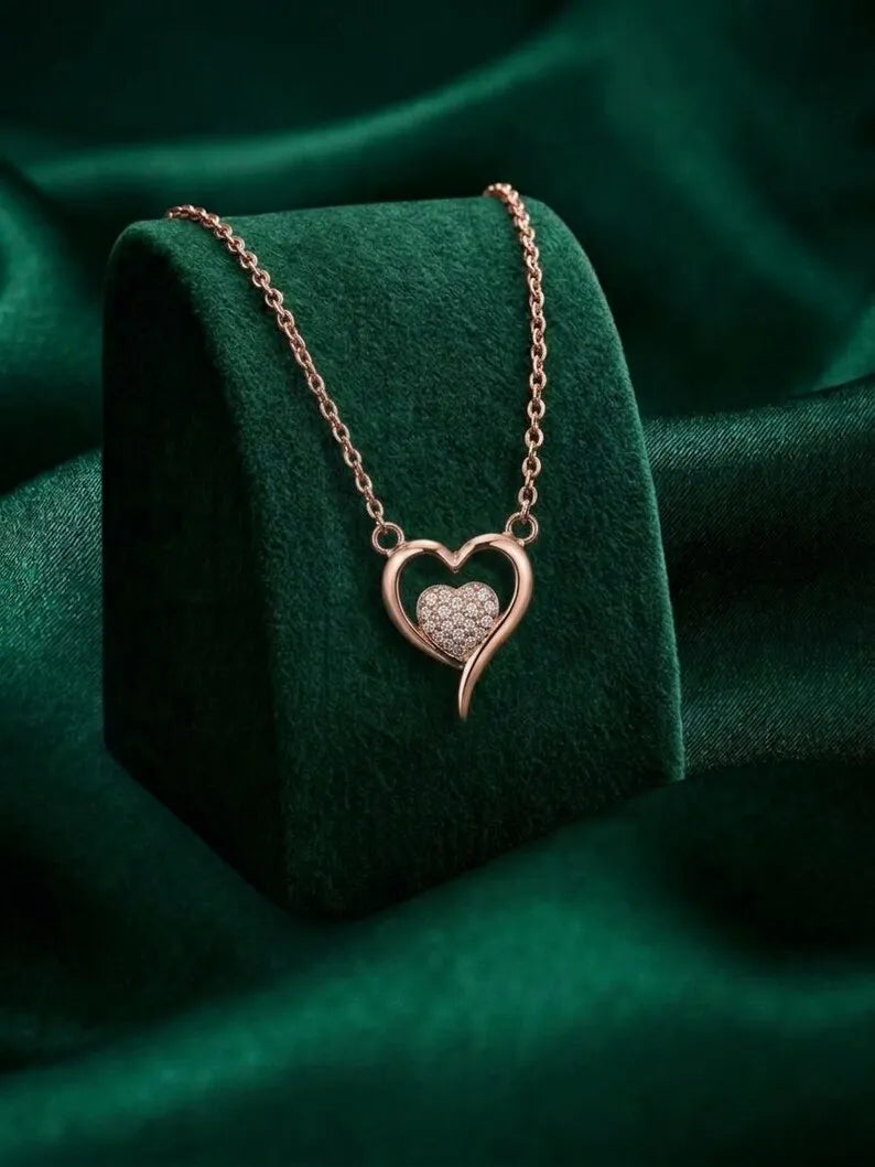 A rose gold heart pendant necklace with a pavé center, displayed on a dark green velvet jewelry stand against a matching green fabric background.