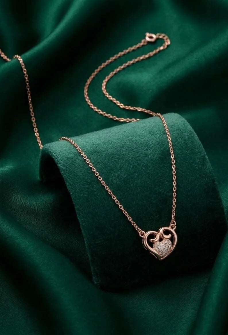 A rose gold heart-shaped pendant necklace with a pavé-set clear stone, displayed on a dark green velvet jewelry stand.