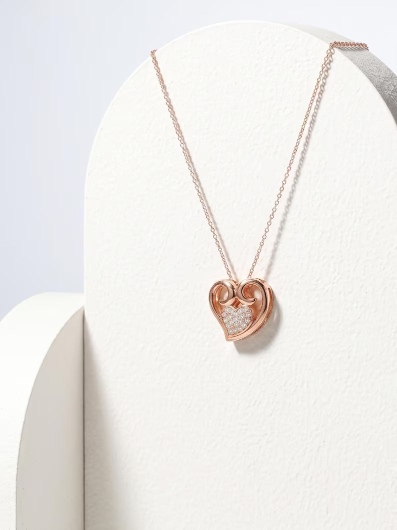 A rose gold heart-shaped pendant necklace with a pavé diamond center, displayed on a white curved jewelry stand.