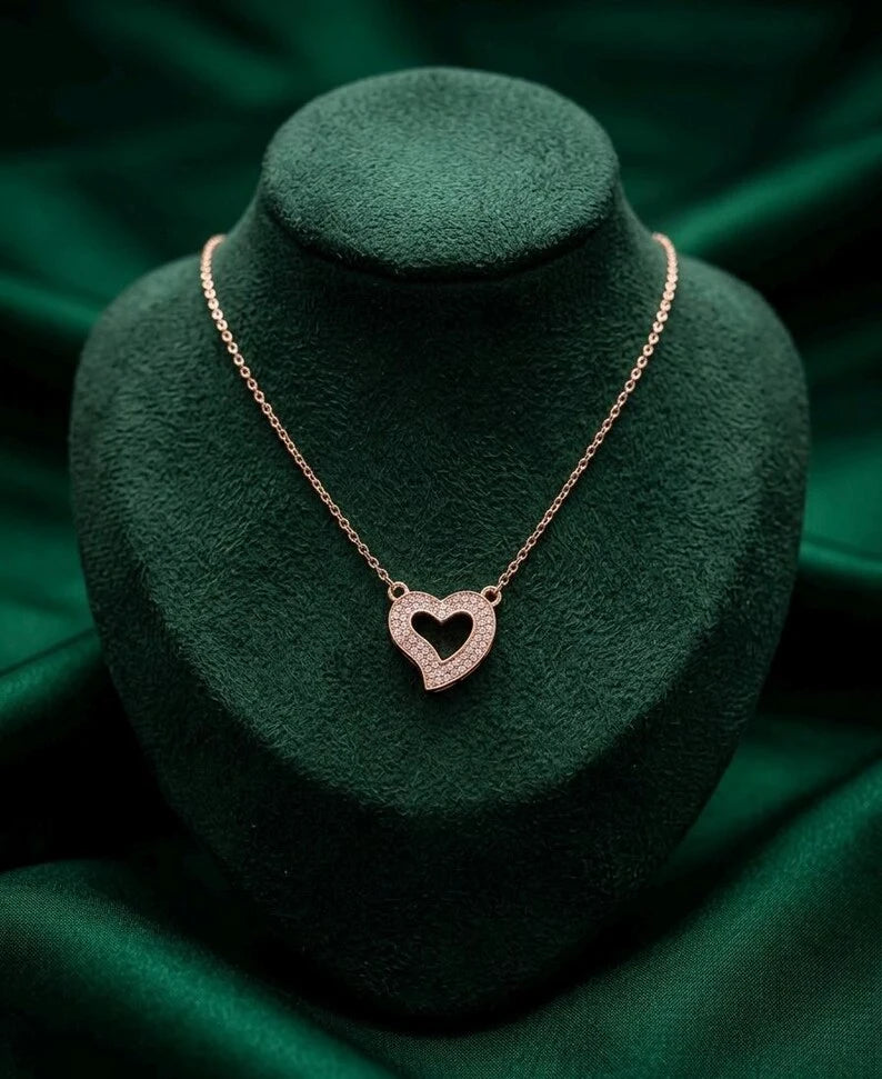 A rose gold heart-shaped pendant necklace displayed on a dark green velvet jewelry bust against a matching green fabric background.