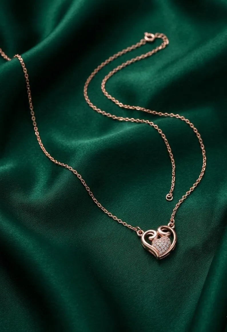 A rose gold heart-shaped pendant necklace with a pavé-set stone center, resting on a draped emerald green satin fabric background.