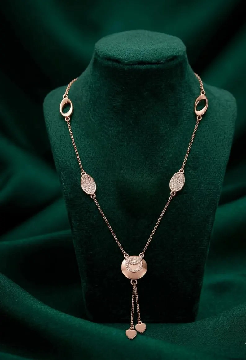 A rose gold heart pendant necklace displayed on a dark green velvet jewelry bust. The necklace features a delicate chain with oval and textured oval links, a central round disc, and two small dangling heart charms at the bottom.
