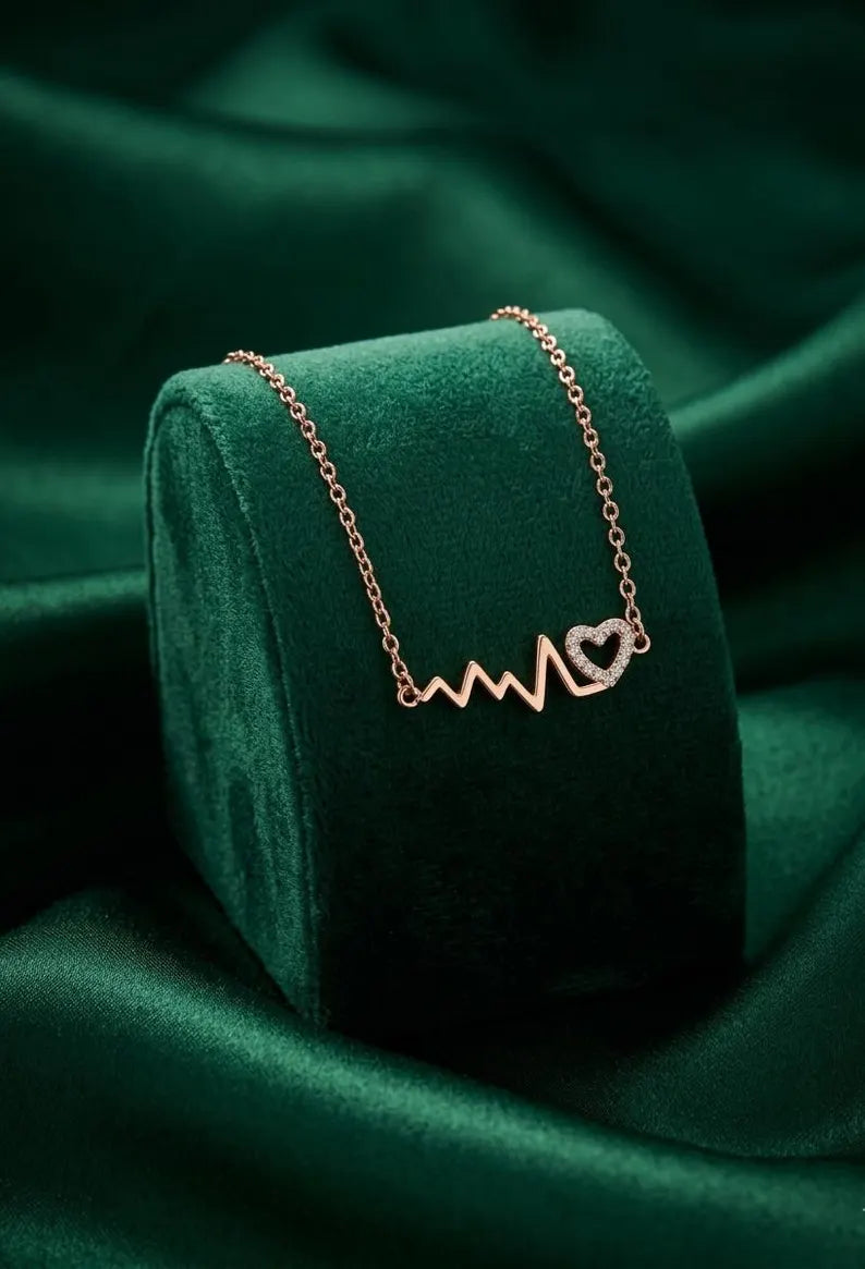 A rose gold necklace with a heartbeat line and heart pendant, displayed on a dark green velvet jewelry stand against a matching green satin background.