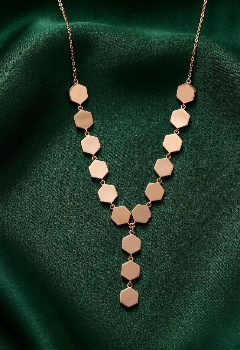 A rose gold Y-necklace with hexagonal disc charms, displayed on a draped dark green satin fabric.