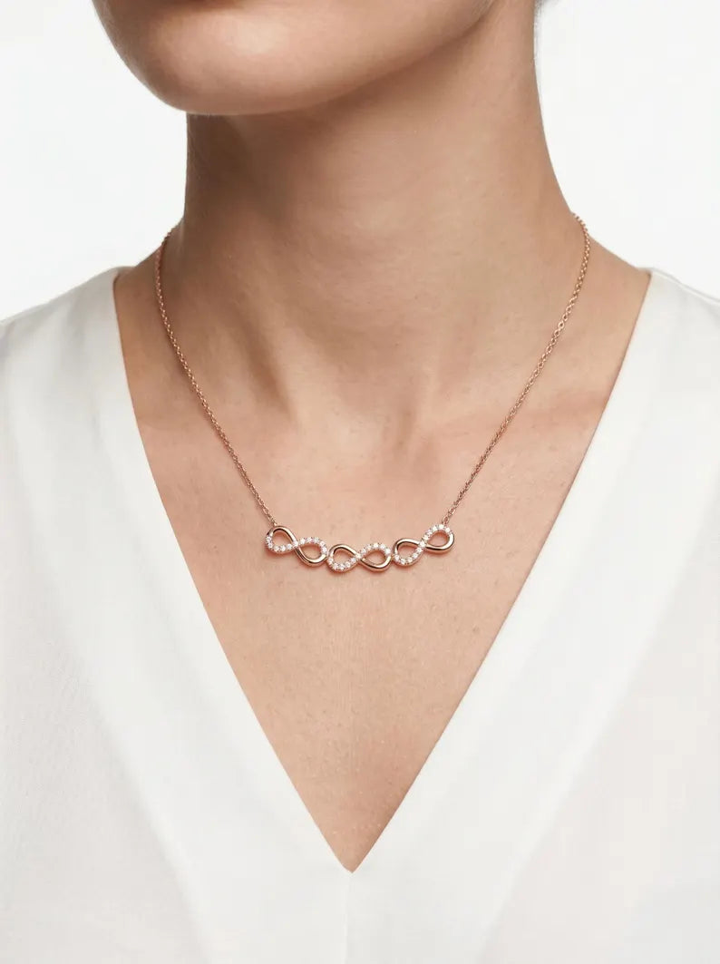 A close-up of a model wearing a delicate rose gold necklace with five interlocking infinity loops, each adorned with small clear crystals, paired with a white V-neck top.