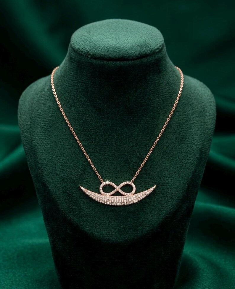 A rose gold necklace with a crescent-shaped infinity pendant paved with small clear stones, displayed on a dark green velvet jewelry bust.