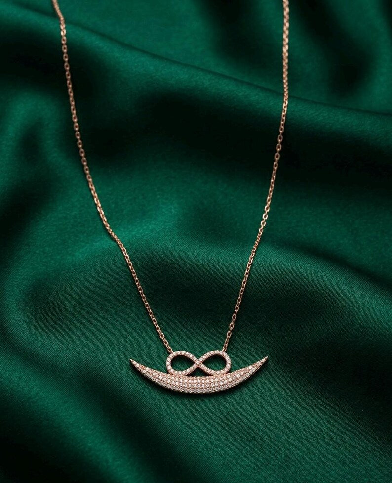 A rose gold necklace featuring a crescent-shaped pendant paved with small clear stones and topped with an infinity symbol, displayed on a draped dark green satin fabric.