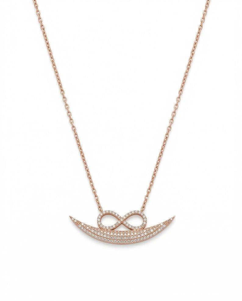 A rose gold necklace featuring a crescent moon and infinity symbol pendant, both adorned with small pavé-set clear stones, shown against a plain white background.