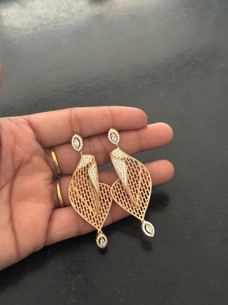 A pair of gold-toned dangle earrings with intricate filigree cutouts and small clear stone accents, held in a person's hand against a dark gray background.