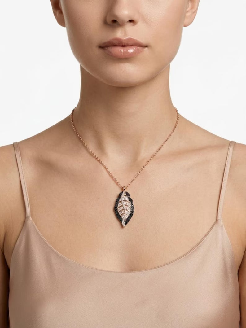 A close-up of a model wearing a delicate rose gold chain necklace with a leaf-shaped pendant featuring black and white textured detailing, paired with a beige satin camisole top against a clean white background.