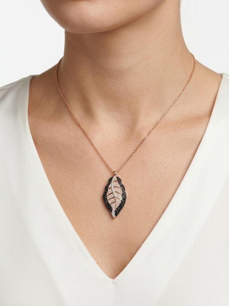 A close-up of a model wearing a delicate rose gold necklace with a leaf-shaped pendant featuring black and white textured detailing, paired with a white V-neck top.
