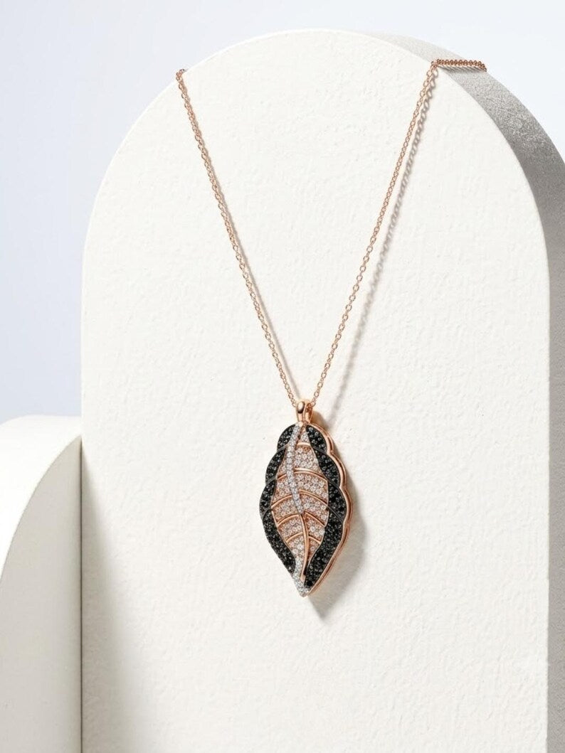 A delicate rose gold chain necklace with a leaf-shaped pendant, displayed on a white curved stand. The pendant features a black textured border and a central design resembling a dried leaf with fine brown and gold lines.