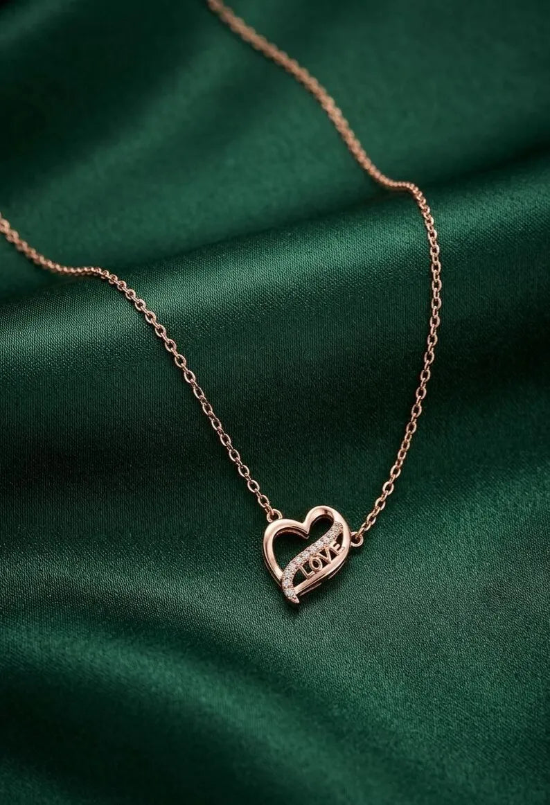 A delicate rose gold chain necklace with a heart-shaped pendant engraved with the word 'LOVE', displayed on a draped dark green satin fabric.