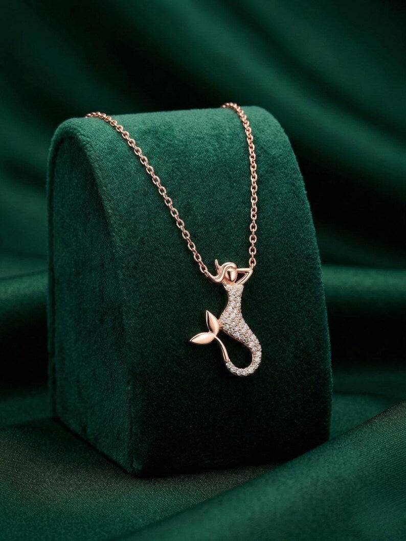 A rose gold mermaid necklace with a pavé-set tail, displayed on a dark green velvet jewelry stand against a matching dark green fabric background.