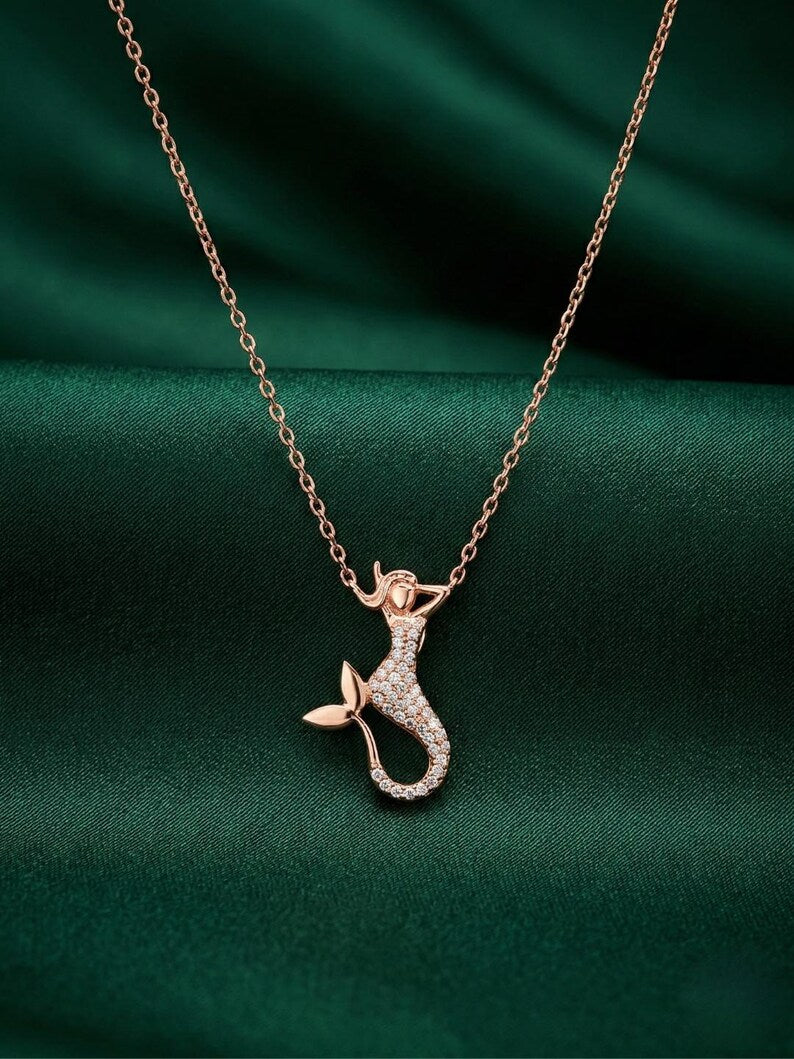 A rose gold mermaid necklace with a small, sparkling pendant, displayed on a dark green satin fabric background.