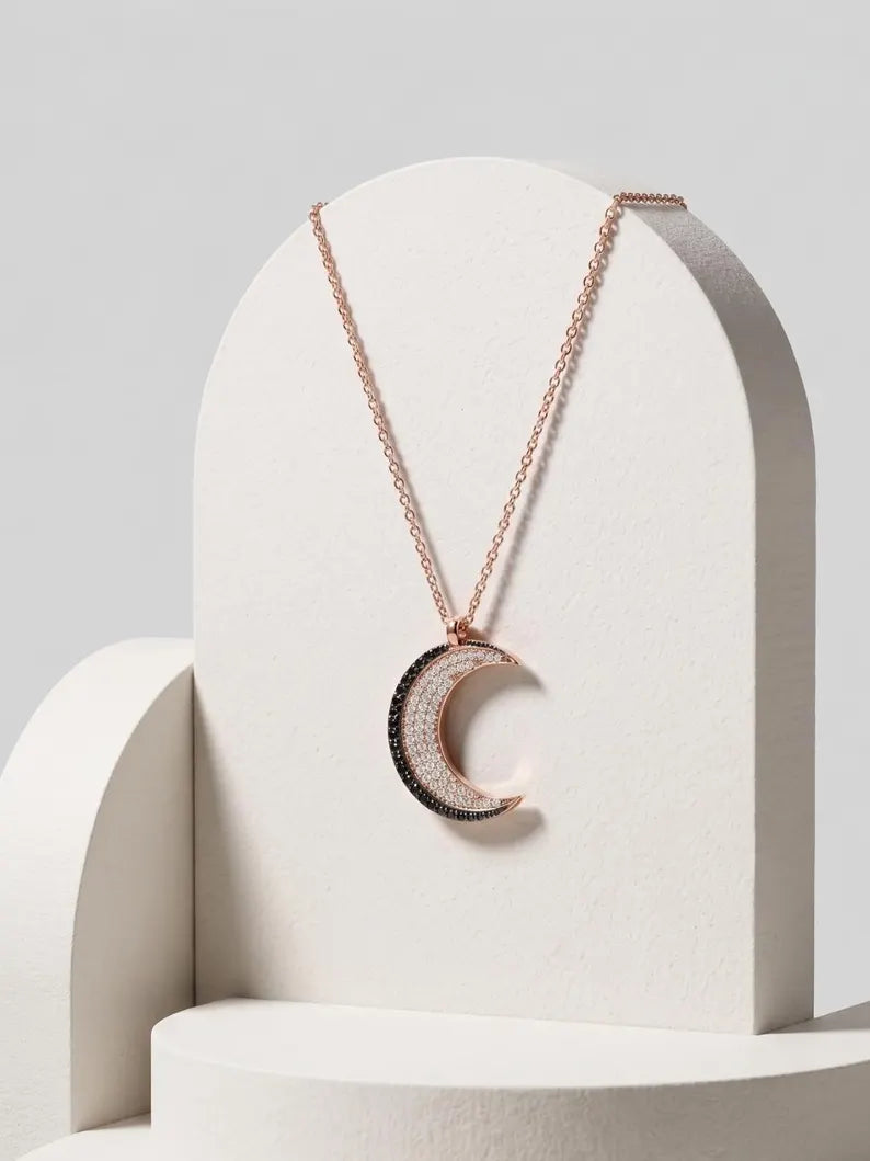 Rose gold crescent moon pendant necklace with a textured black outer ring, displayed on a white geometric stand against a light gray background.