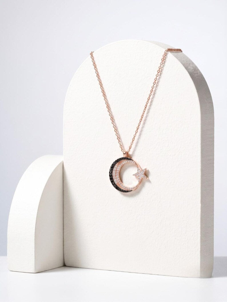 A rose gold necklace with a crescent moon and star pendant, displayed on a white arched stand against a light gray background.