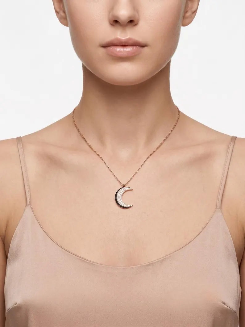 A close-up of a model wearing a delicate rose gold crescent moon pendant necklace with a black inlay, styled with a beige satin camisole top against a light gray background.