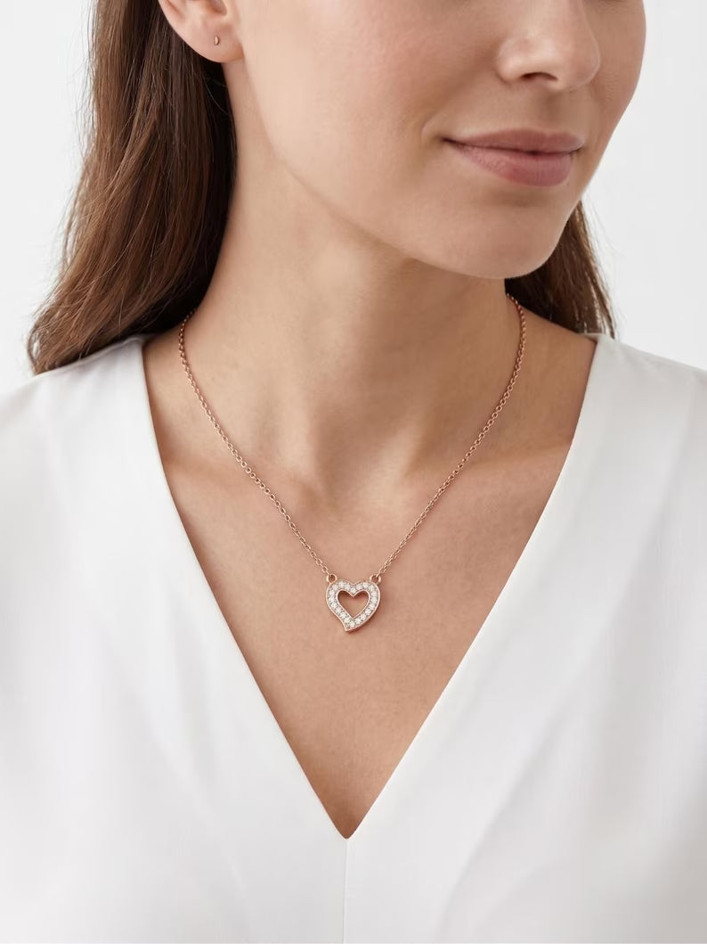 A close-up of a woman wearing a delicate rose gold heart-shaped pendant necklace with a pavé-set clear stone border, paired with a white V-neck top.