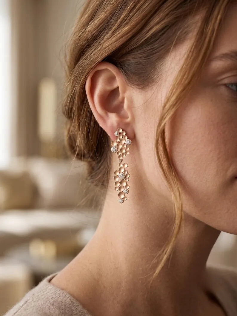 A close-up of a woman's ear and side profile wearing a delicate rose gold dangle earring adorned with small clear crystals and clustered floral details.