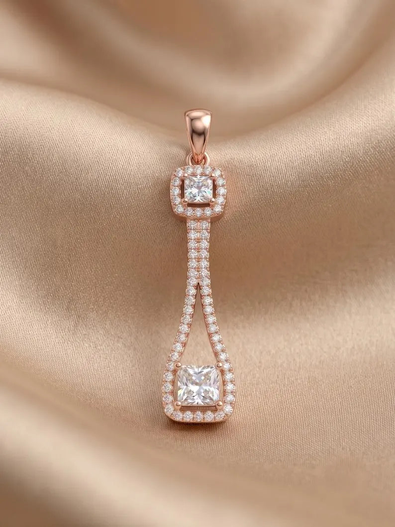 A rose gold pendant with a teardrop-shaped pavé setting and square cushion-cut diamond accents, displayed on a draped beige satin fabric.
