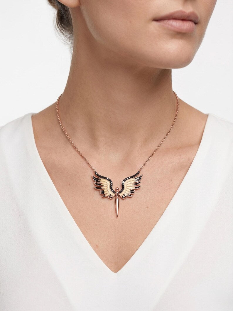 A close-up of a model wearing a rose gold wing pendant necklace with black enamel details, paired with a white V-neck top against a clean white background.