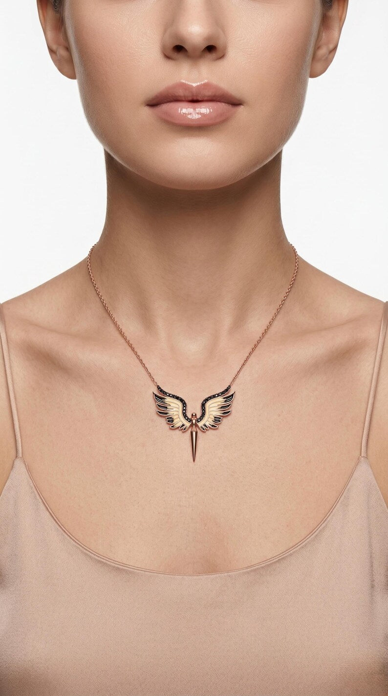 A close-up of a model wearing a delicate rose gold necklace with a winged dagger pendant, styled with a beige camisole against a clean white background.
