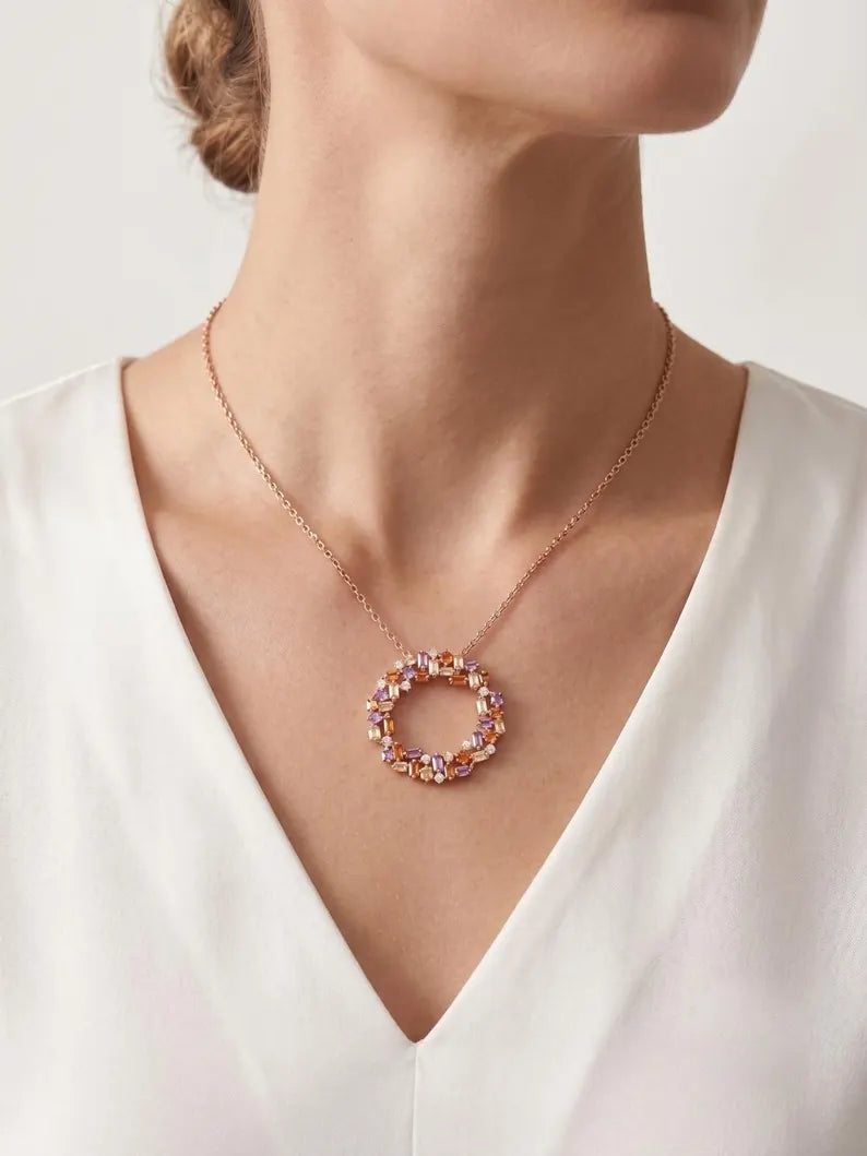 A close-up of a model wearing a delicate rose gold necklace with a circular pendant adorned with small pink and purple crystals, paired with a white V-neck top.