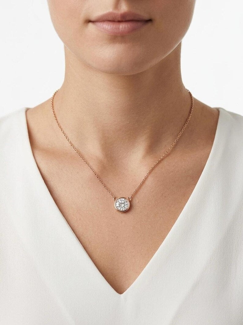 A close-up of a model wearing a delicate rose gold chain necklace with a round bezel-set clear gemstone pendant, styled with a white V-neck top.