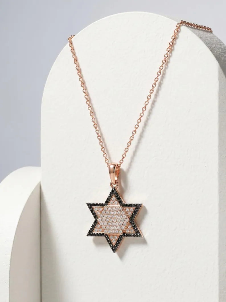 A rose gold Star of David pendant necklace with a textured center and black outer border, displayed on a white curved stand.