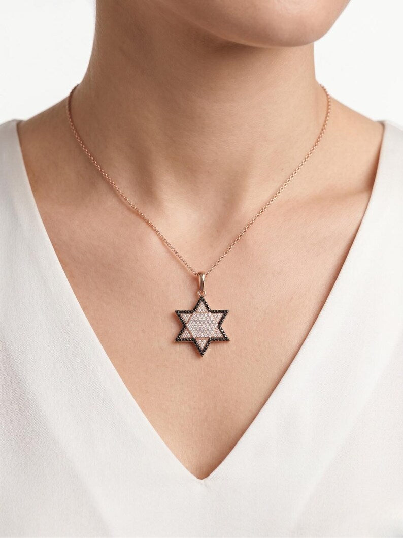 A close-up of a model wearing a delicate rose gold chain necklace with a Star of David pendant. The pendant features a textured, light-colored inlay with a dark rose gold border, set against a clean white background.
